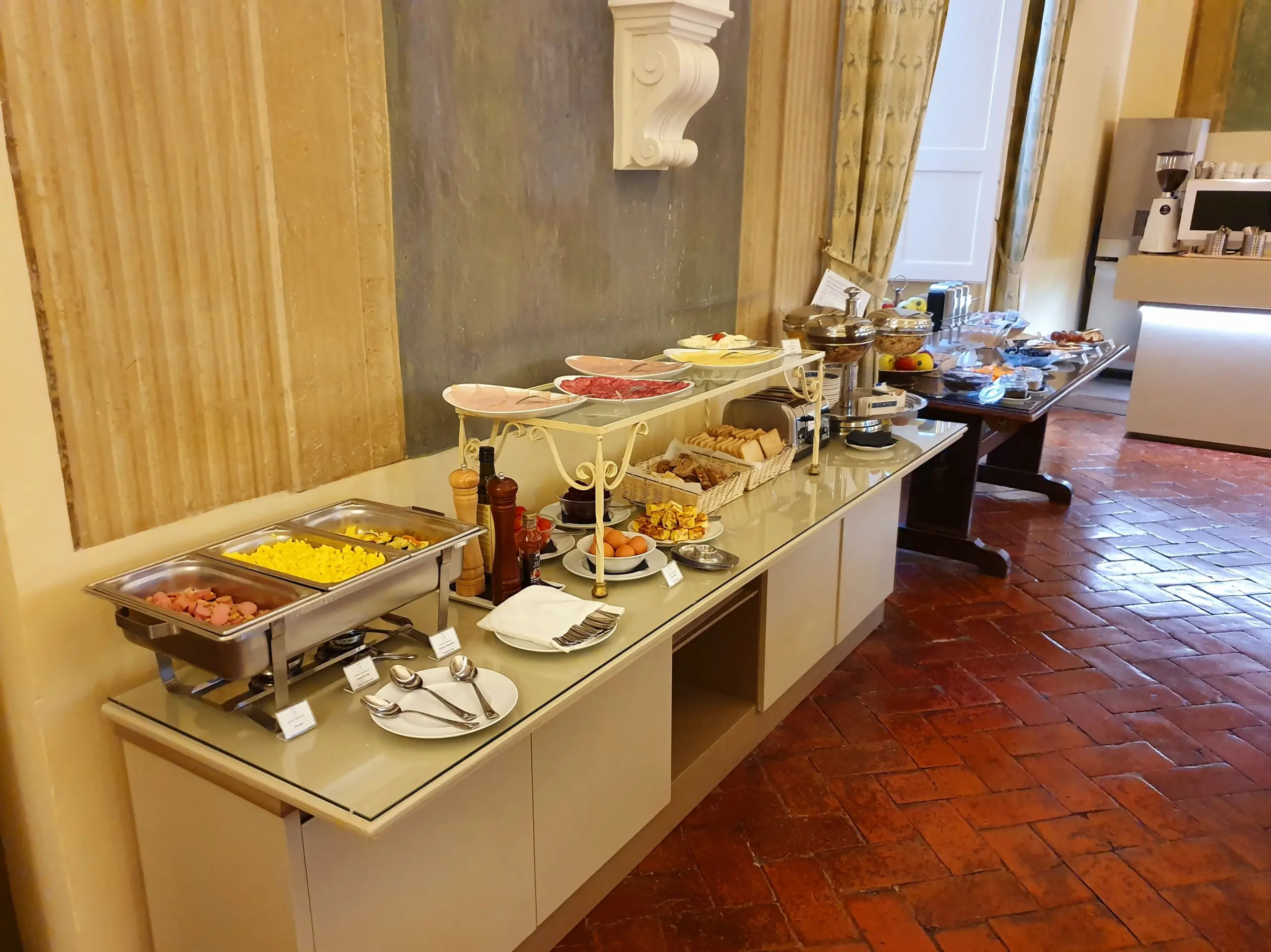 Buffet della colazione con piatti caldi, salumi, pane tostato e frutta lungo la parete della sala colazione